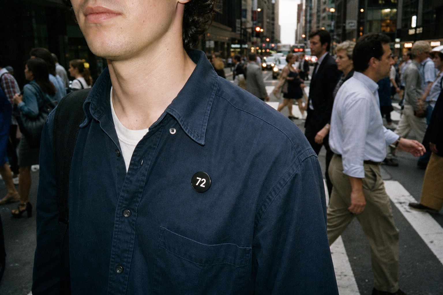 Young adult wearing an ultra-thin black NUMBR badge displaying only 72 on a shirt in a crowded city crosswalk